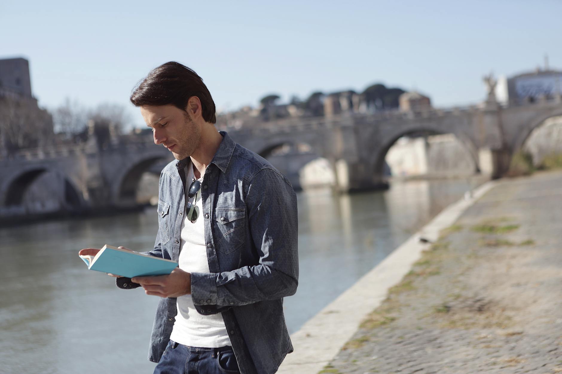 young man in denim jacket is reading a book near river side